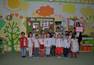 Dzieci z grupy IV pozują do wspólnego zdjęcia. Dzieci są we większości przebrane w stroje w polskich kolorach narodowych. Razem z dziećmi stoi nauczycielka.